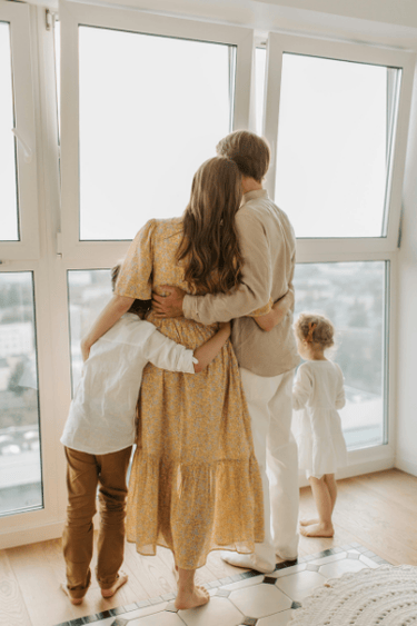 A family standing together by a large window, looking outside while discussing household finances.
