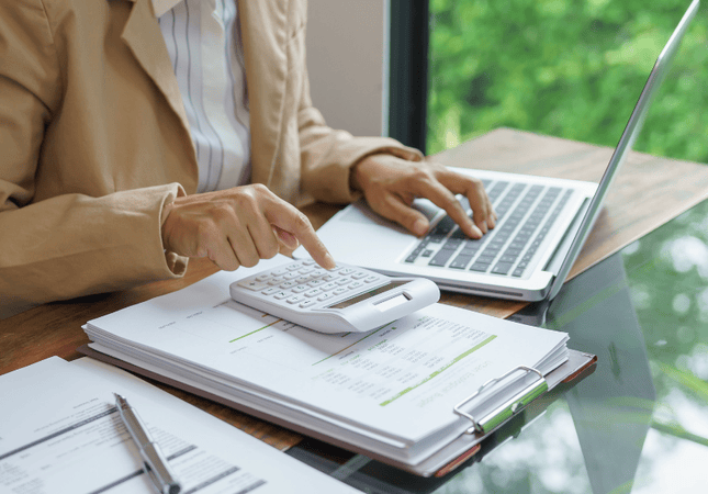 Woman using a calculator and laptop while reviewing household finances.